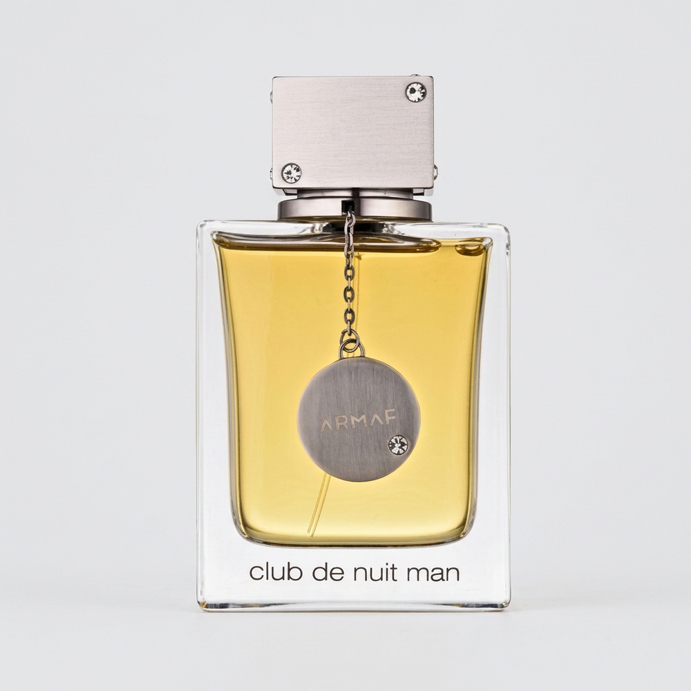 A bottle of Club de Nuit Man fragrance by Armaf, featuring a clear glass bottle filled with golden-yellow liquid, a silver-toned circular hanging medallion, and a white rectangular cap, set against a neutral gray background.