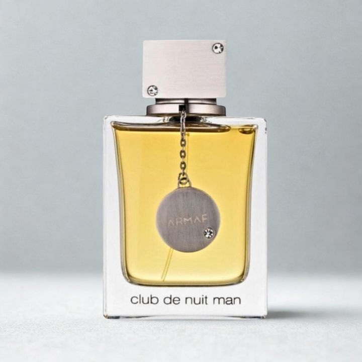 A bottle of Club de Nuit Man fragrance by Armaf, featuring a clear glass bottle filled with golden-yellow liquid, a silver-toned circular hanging medallion, and a white rectangular cap, set against a neutral gray background.