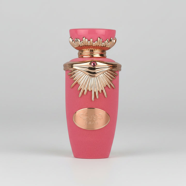 A pink perfume bottle with ornate gold-tone detailing, including a sunburst pattern and a small red gem accent, on a plain white background.
