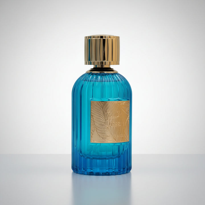 A blue ribbed glass perfume bottle with a gold metallic cap and a gold rectangular label featuring an embossed palm leaf design, set against a plain white background.